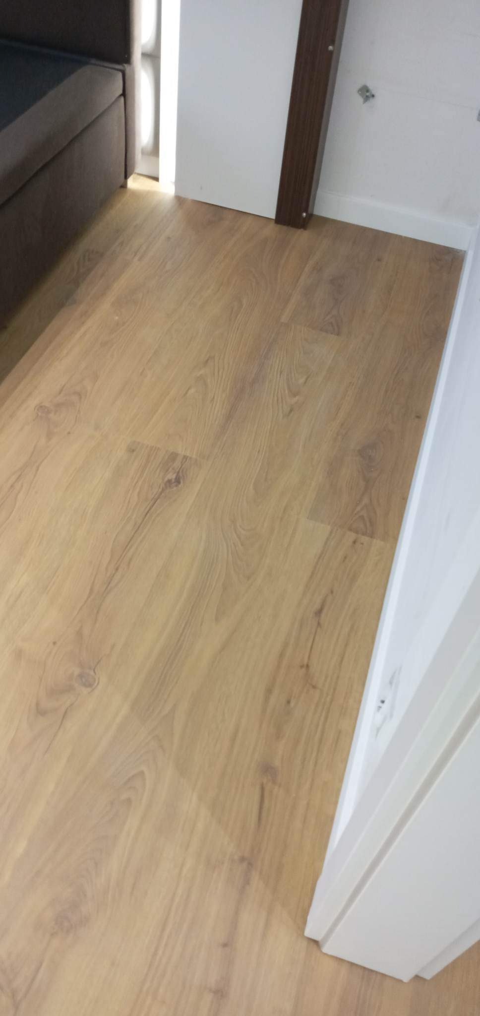 Nowo ułożony parkiet laminowany w jasnym odcieniu dębu w rogu pokoju, widoczny fragment sofy i biała listwa przypodłogowa.