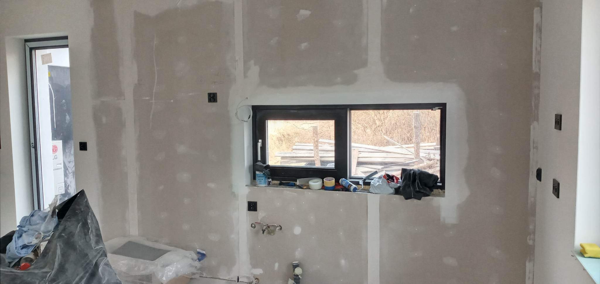 Wnętrze w trakcie prac wykończeniowych: ściany pokryte gładzią gipsową, widoczne puszki elektryczne, okno z widokiem na składowane drewno na zewnątrz, narzędzia i materiały budowlane na parapecie...