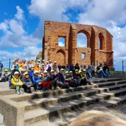 Grupa dzieci w żółtych czapkach siedzi na schodach przed ruinami ceglanego budynku pod błękitnym niebem z chmurami, prawdopodobnie podczas wycieczki.