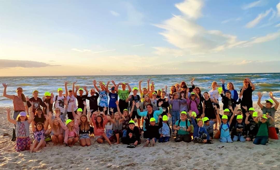 Grupa dzieci w neonowych czapkach pozuje na plaży w Sosnowcu, z morzem i zachodzącym słońcem w tle.