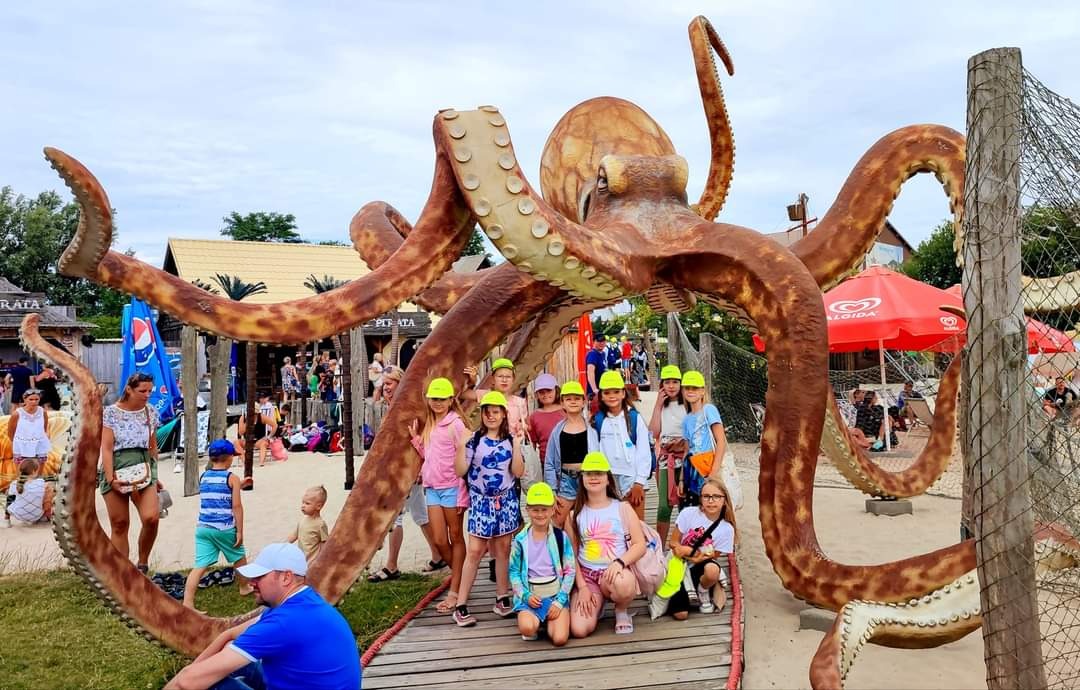 Grupa dzieci w neonowych czapkach pozuje na tle gigantycznej, brązowej rzeźby ośmiornicy na piaszczystym placu zabaw z budynkami w tle.