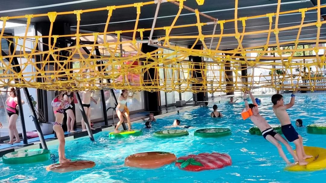 Kryty basen z przeszkodami: dzieci bawią się na torze linowym zawieszonym nad wodą i na pływających platformach w kształcie jedzenia.