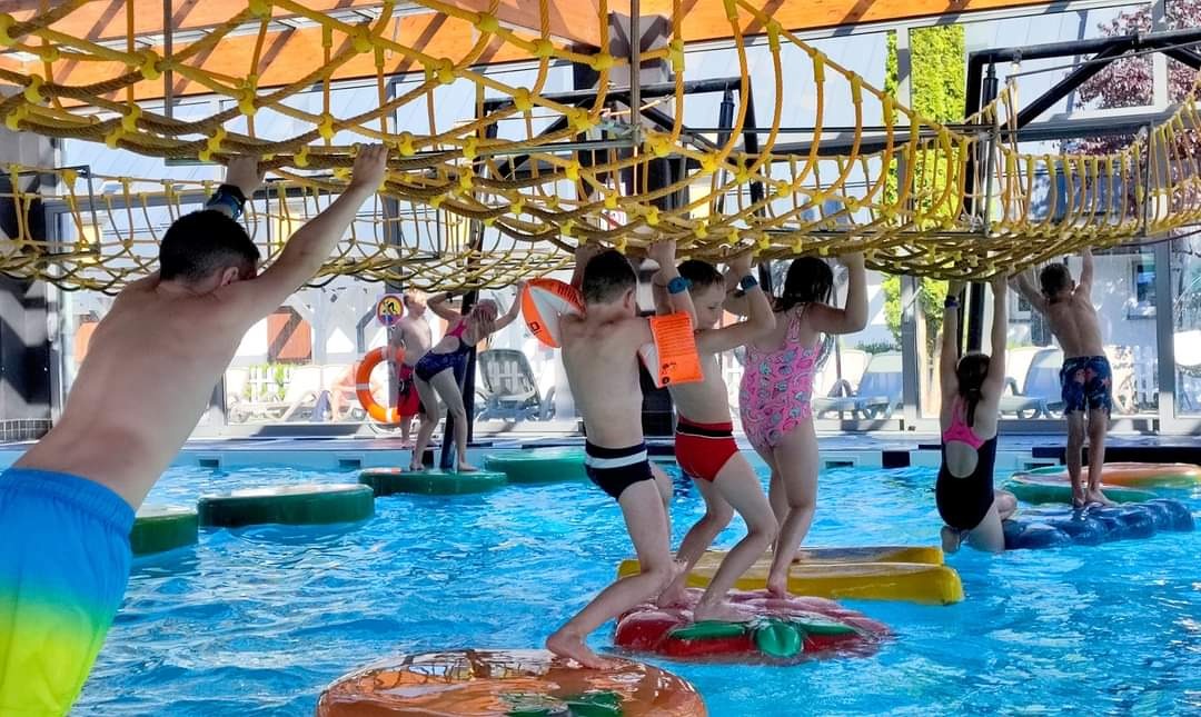 Dzieci w różnokolorowych strojach kąpielowych przechodzą przez wodny tor przeszkód z siatką linową zawieszoną nad głowami, niektóre z kołami ratunkowymi, w jasnym, przestronnym wnętrzu basenu...