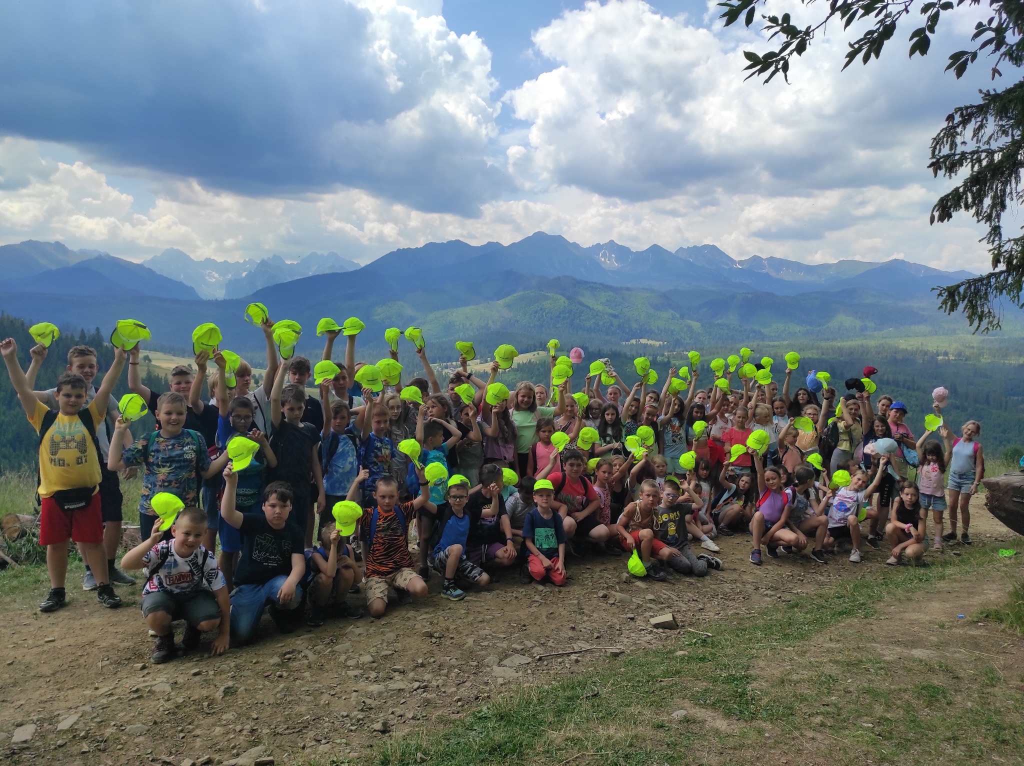 Duża grupa dzieci w neonowych żółtych czapkach pozuje na tle górskiego krajobrazu z widocznymi szczytami i chmurami.