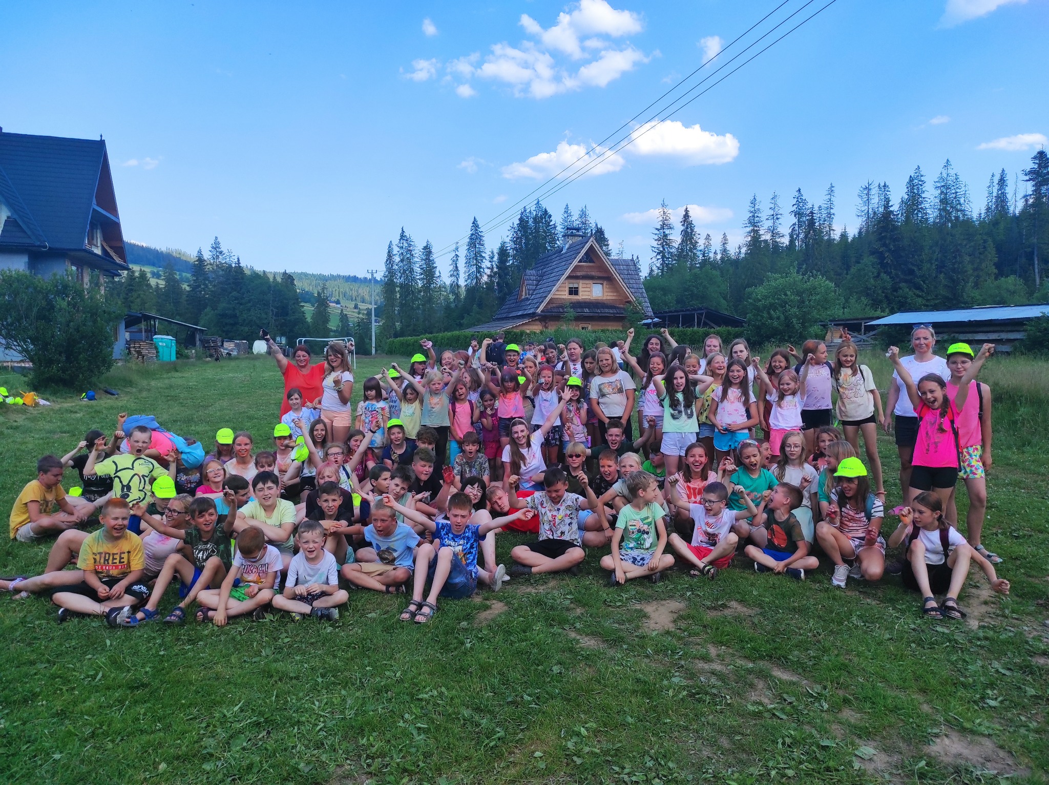 Duża grupa uśmiechniętych dzieci siedzi na trawie przed drewnianym domem w górach, część z nich ma podniesione ręce w geście radości, a niektórzy noszą neonowe czapki.