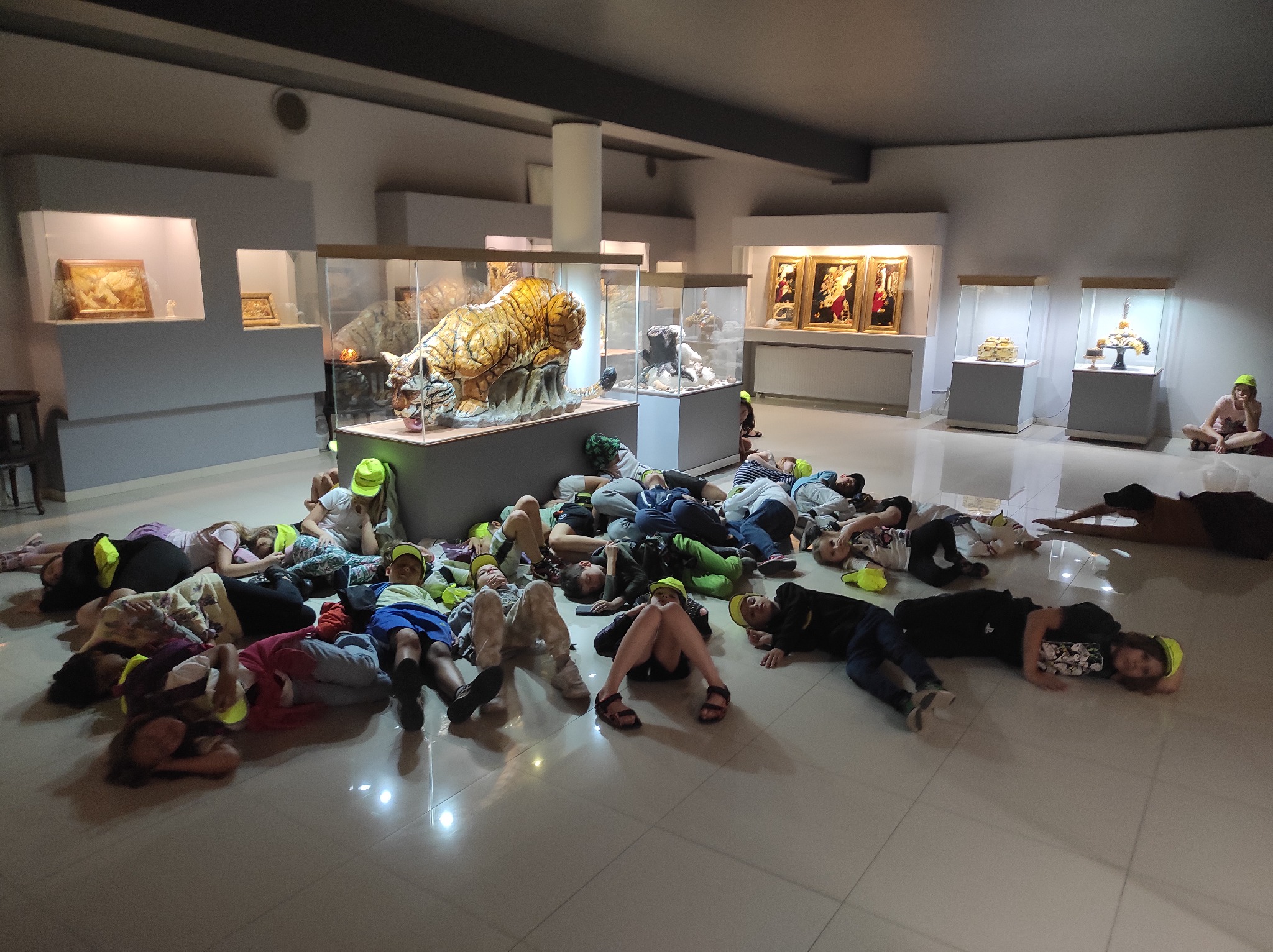 Grupa dzieci w neonowych czapkach leży na podłodze w muzeum, otoczona szklanymi gablotami z eksponatami.