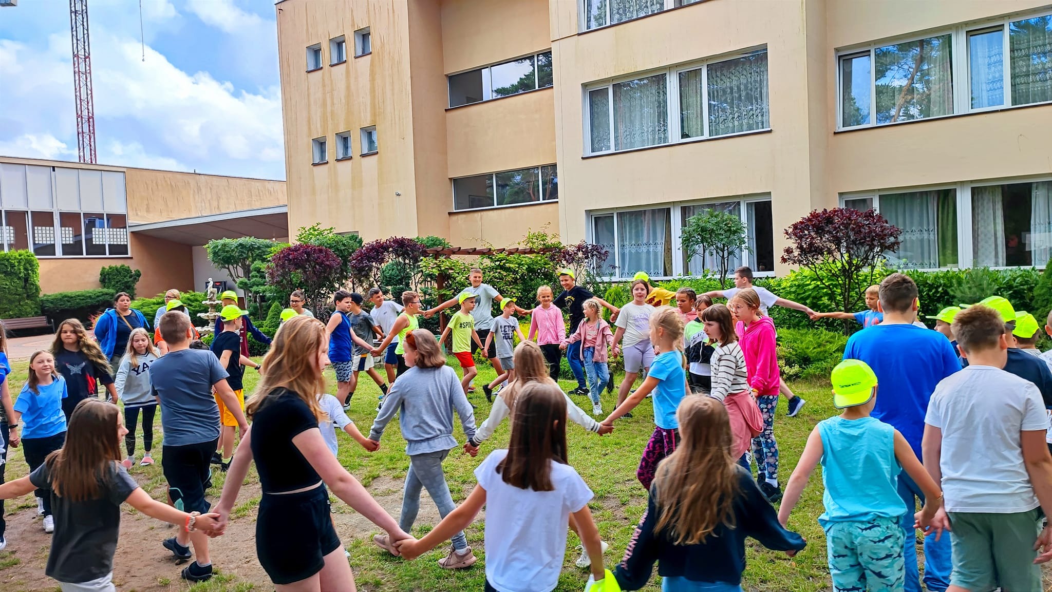 Grupa dzieci w neonowych czapkach trzymająca się za ręce, tworząca koło na trawniku przed budynkiem z jasną elewacją i dużymi oknami.