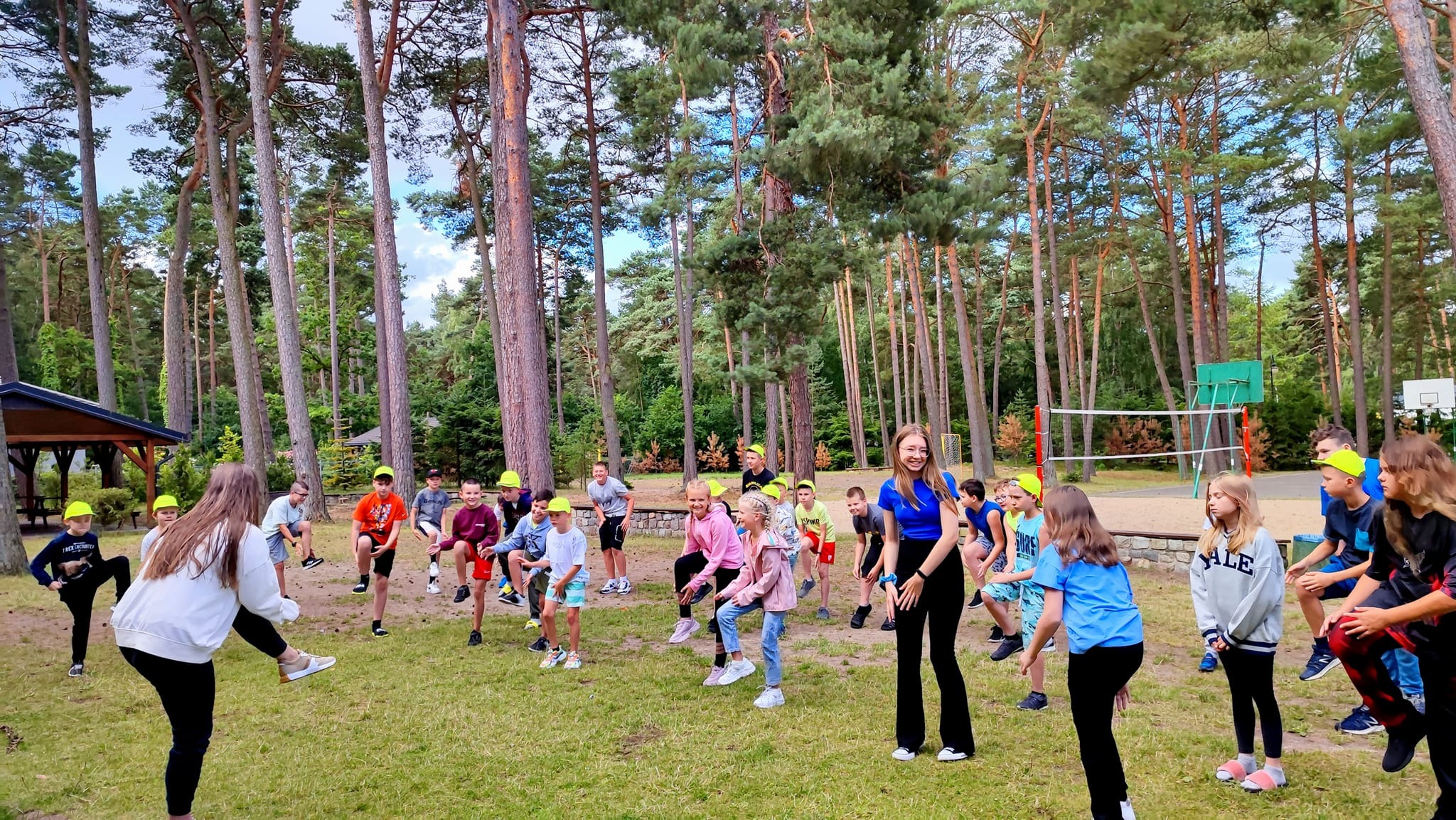 Grupa dzieci i młodzieży w żółtych czapkach ćwiczy na trawie otoczonej wysokimi sosnami. W tle widoczny plac zabaw z siatką do siatkówki i koszem do koszykówki.