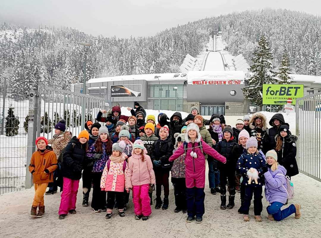 Grupa dzieci w zimowych ubraniach pozuje na tle Wielkiej Krokwi w Zakopanem, widoczny śnieg i budynki.