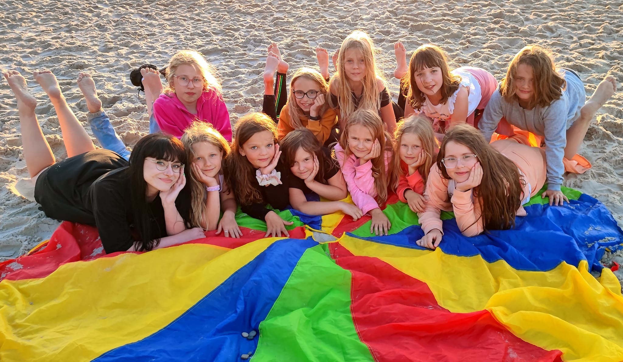 Grupa uśmiechniętych dziewcząt i jedna dorosła kobieta leżą na piasku na kolorowej chuście animacyjnej podczas zajęć rekreacyjnych na plaży, niektóre z nich podpierają głowy rękami, a inne trzymają...