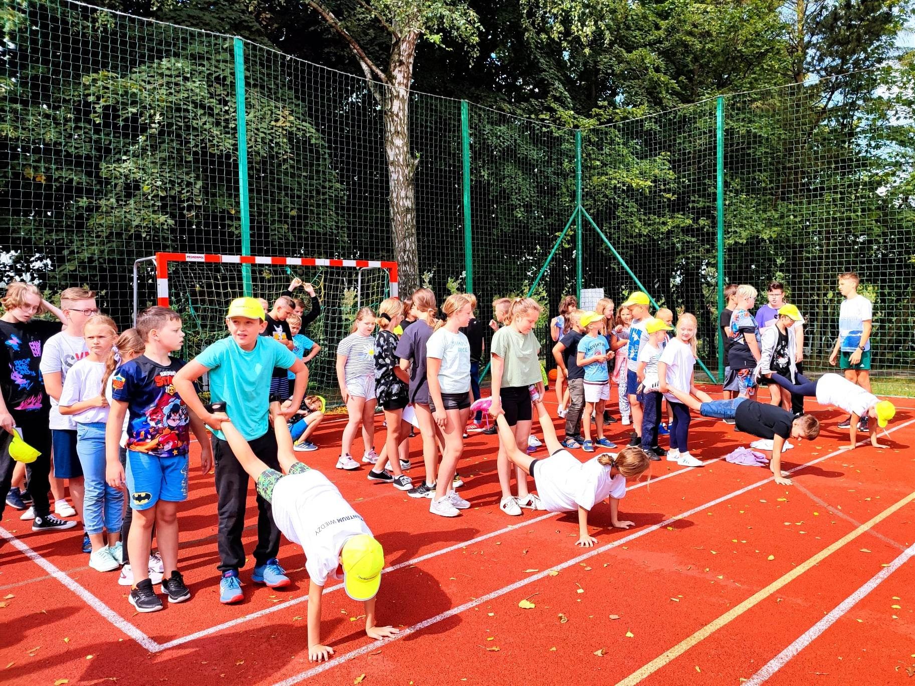 Grupa dzieci w żółtych czapkach na czerwonym boisku sportowym wykonuje ćwiczenia gimnastyczne, w tle widoczna siatka i drzewa.