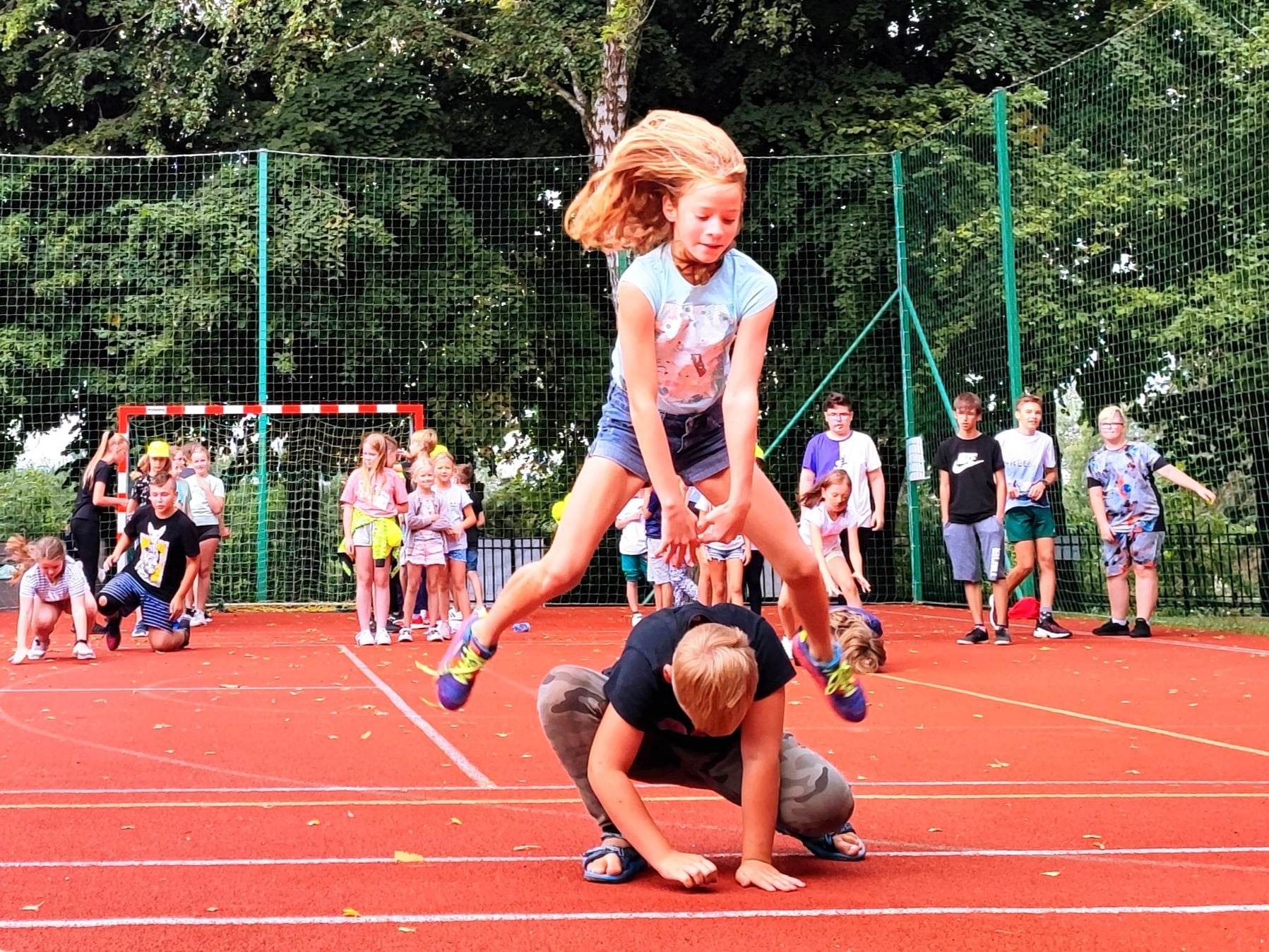 Dzieci skaczące przez siebie na czerwonym boisku podczas zajęć kolonijnych, widoczna siatka i drzewa w tle.