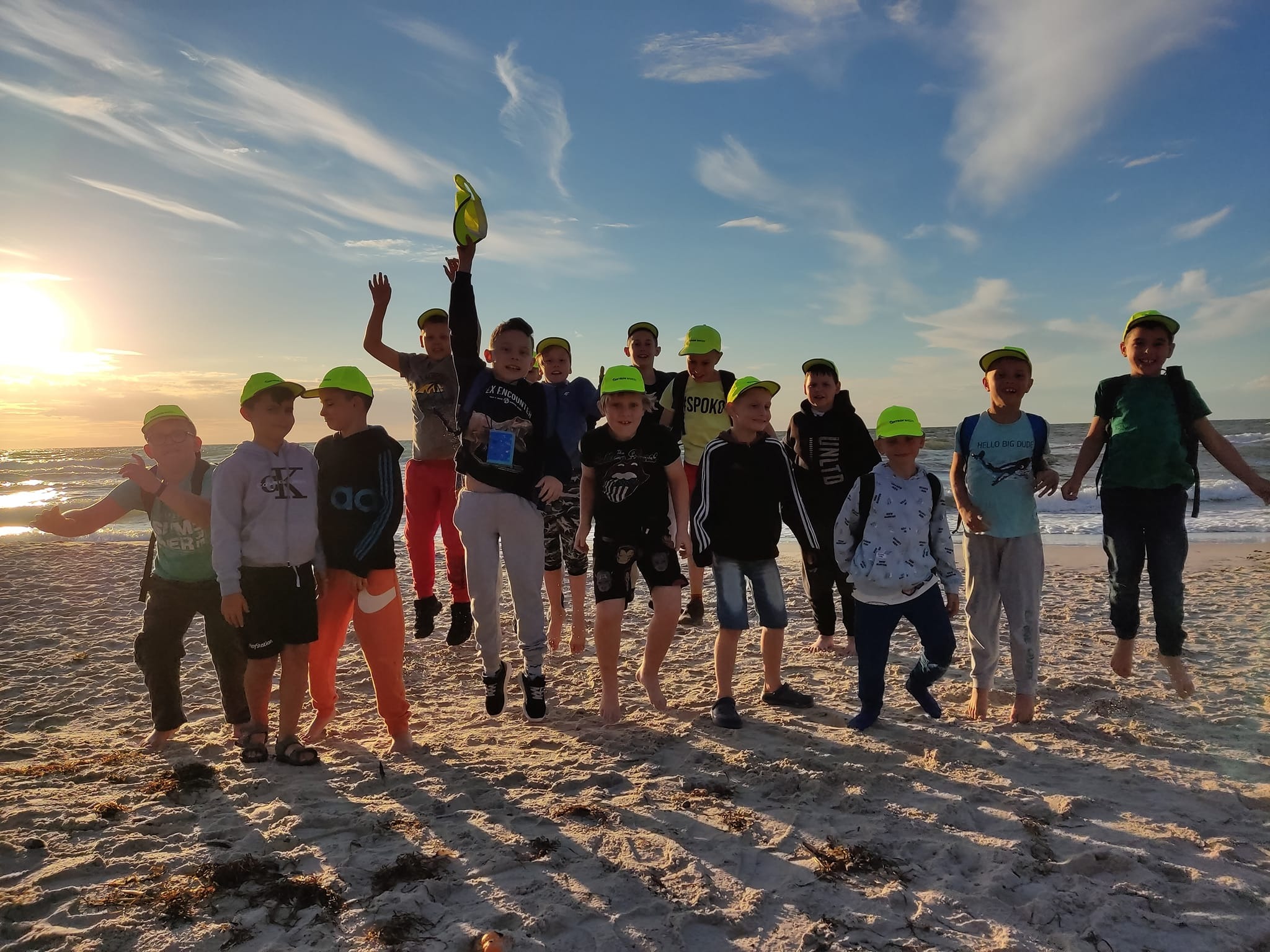 Grupa uśmiechniętych dzieci w neonowych, zielonych czapkach skacze radośnie na piaszczystej plaży o zachodzie słońca, z widocznymi falami morskimi w tle.