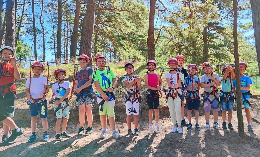 Grupa dzieci w kaskach i uprzężach, stojących w lesie przed linami parku linowego, gotowych do zabawy.