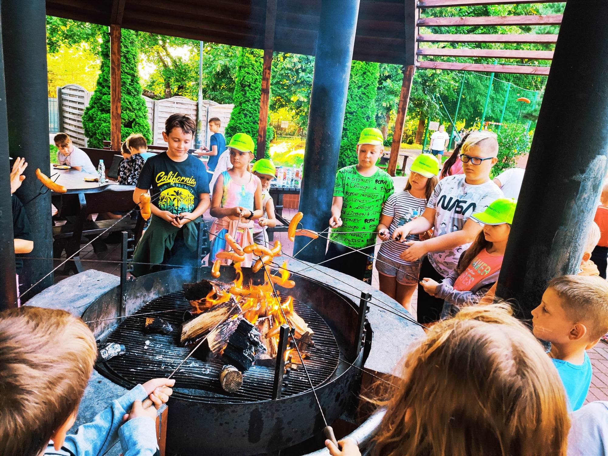 Grupa dzieci w zielonych czapkach zgromadzona wokół paleniska, piekąca kiełbaski na długich prętach, z widocznymi płomieniami i dymem, w tle zieleń drzew i budynki.