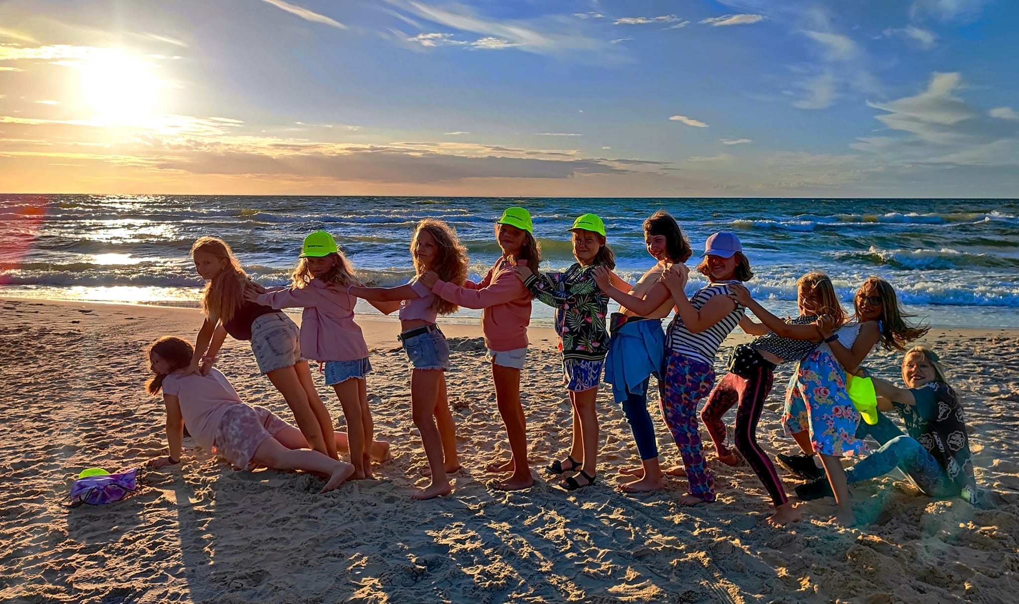 Grupa dzieci w neonowych kapeluszach ustawiona rzędem na plaży, tworząca ludzki tunel przy zachodzącym słońcu nad morzem.