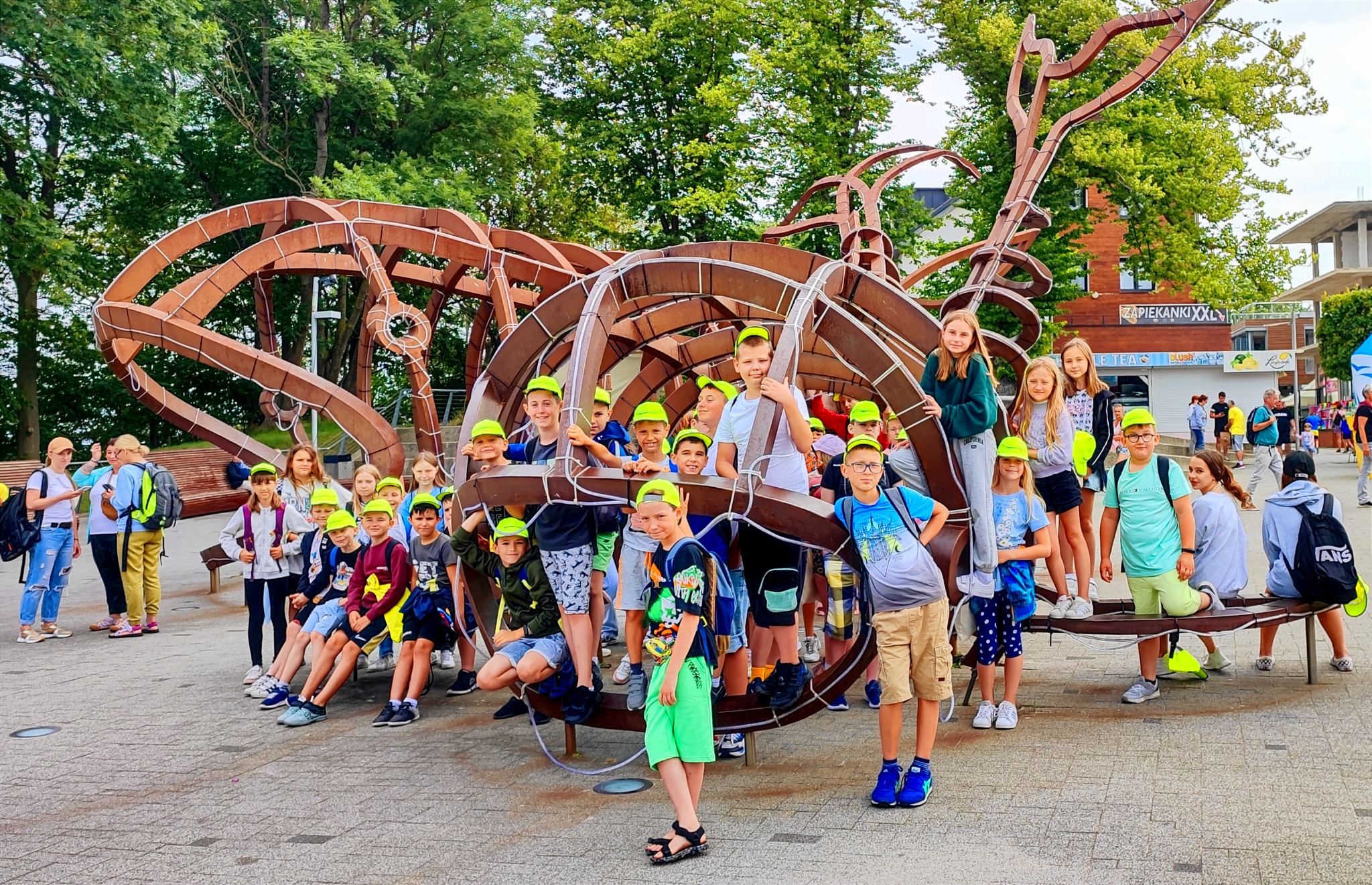 Grupa dzieci w neonowych czapkach pozuje na tle rzeźby przypominającej kosmiczny statek podczas wycieczki.