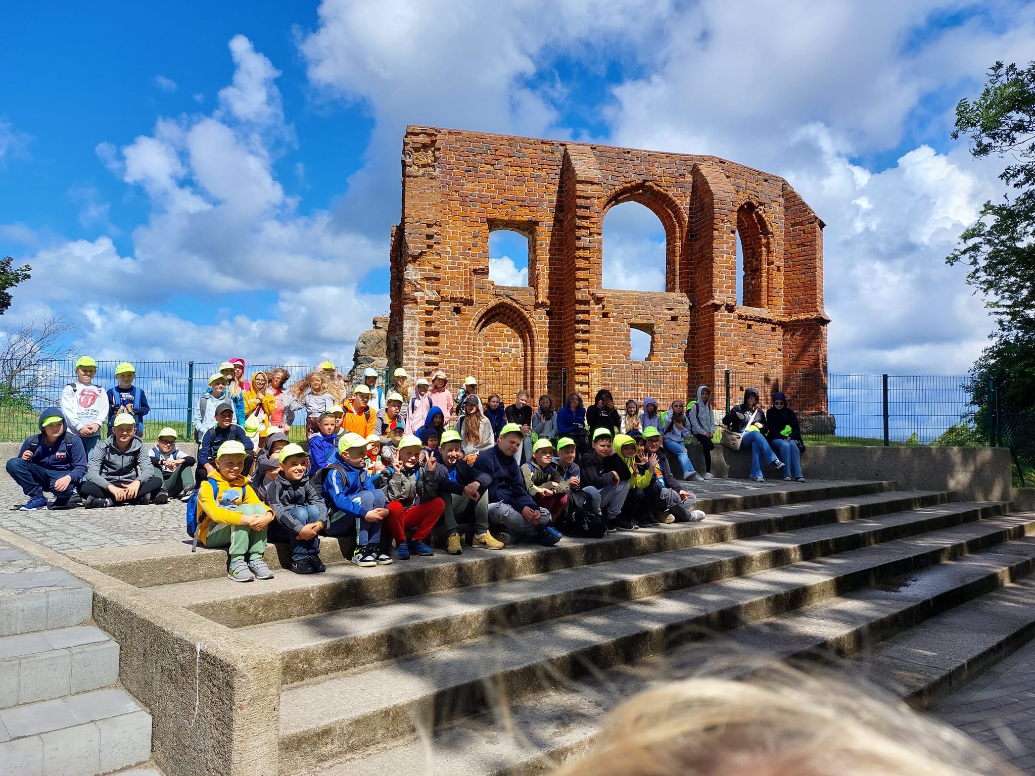 Grupa dzieci w żółtych czapkach siedzi na schodach przed ruinami ceglanego budynku pod błękitnym niebem z chmurami, prawdopodobnie podczas wycieczki.