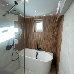 Nowoczesna łazienka z wolnostojącą białą wanną, czarną armaturą i ścianą wykończoną drewnianymi panelami, widok z kabiny prysznicowej, oświetlenie punktowe.