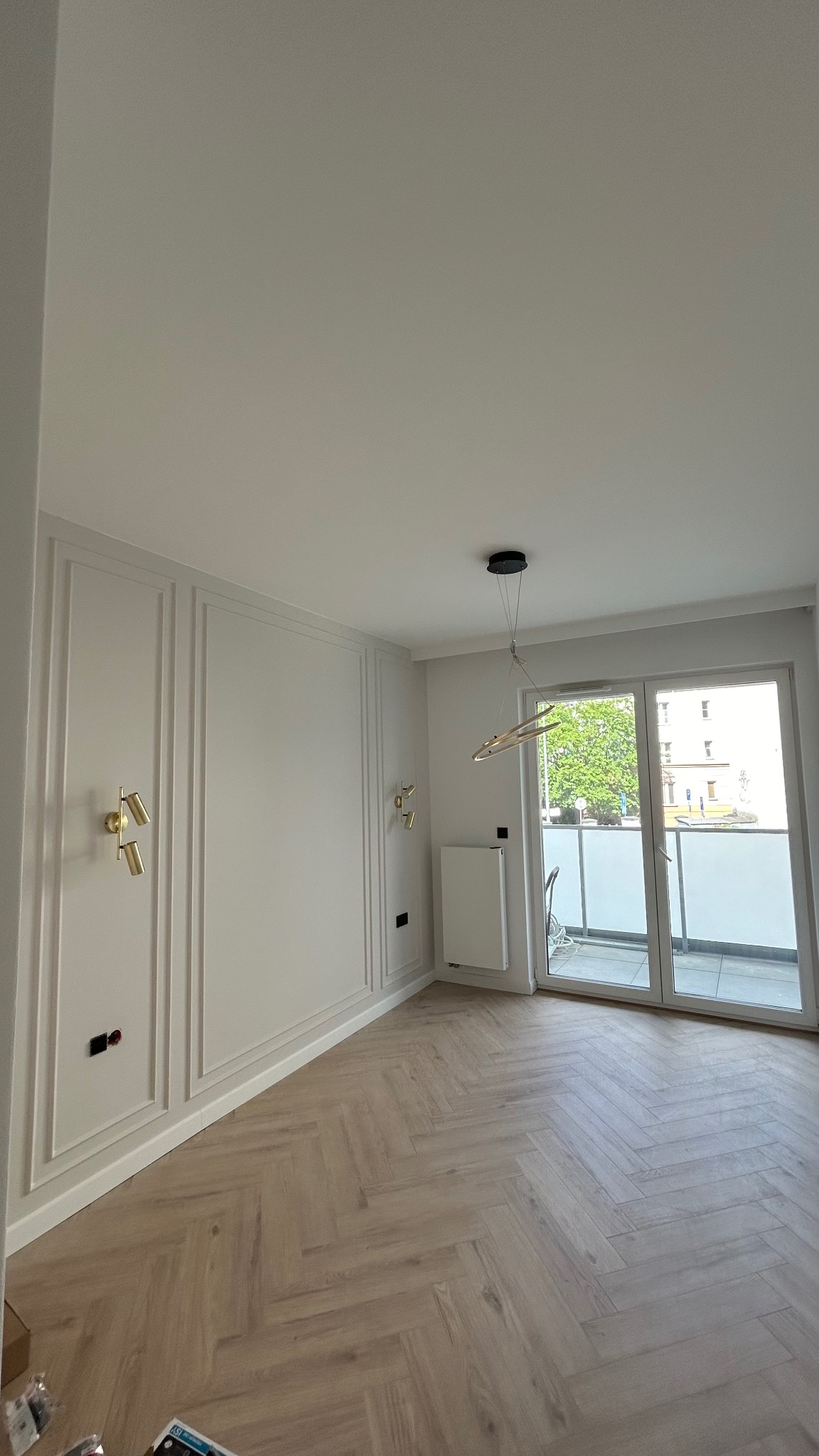 Jasne, przestronne wnętrze z parkietem w jodełkę, ozdobnymi panelami ściennymi, złotymi kinkietami i nowoczesną lampą wiszącą, balkon z widokiem na zieleń.