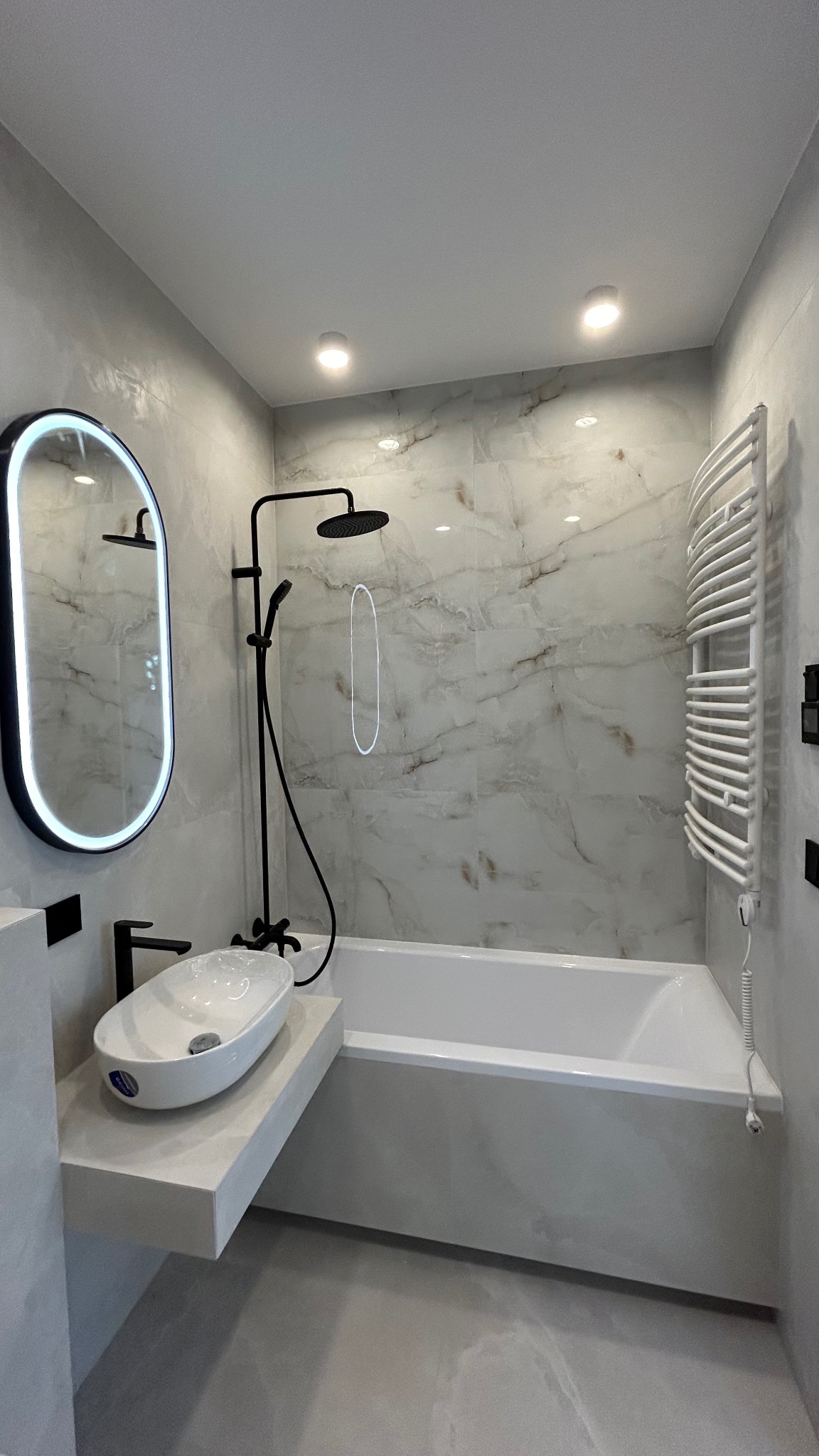Nowoczesna łazienka z owalnym lustrem z podświetleniem LED, białą umywalką nablatową na szarym blacie, czarną armaturą i grzejnikiem drabinkowym. Ściany wykończone płytkami imitującymi marmur.