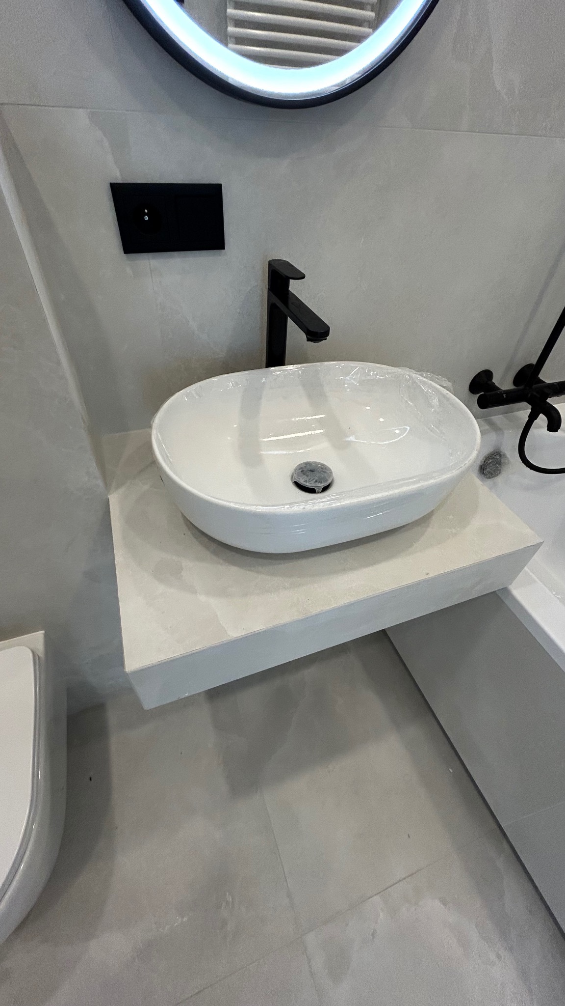 Nowoczesna łazienka w szarościach: owalna umywalka nablatowa z czarną armaturą, podświetlane okrągłe lustro i minimalistyczna spłuczka podtynkowa.