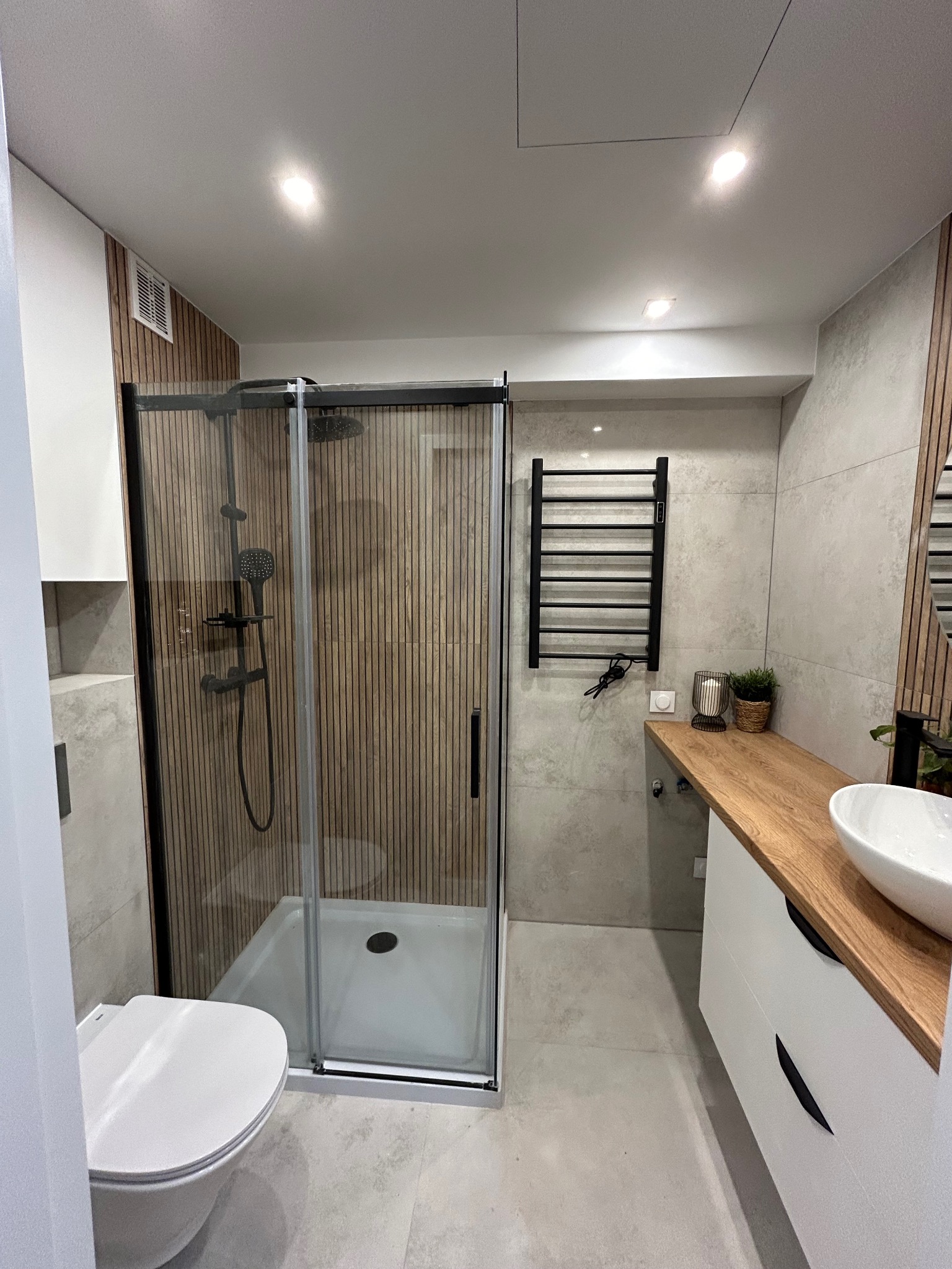 Nowoczesna łazienka z kabiną prysznicową typu walk-in z czarnymi ramami, drewnianymi panelami na ścianie kabiny, umywalką nablatową na drewnianym blacie i czarnym grzejnikiem drabinkowym.