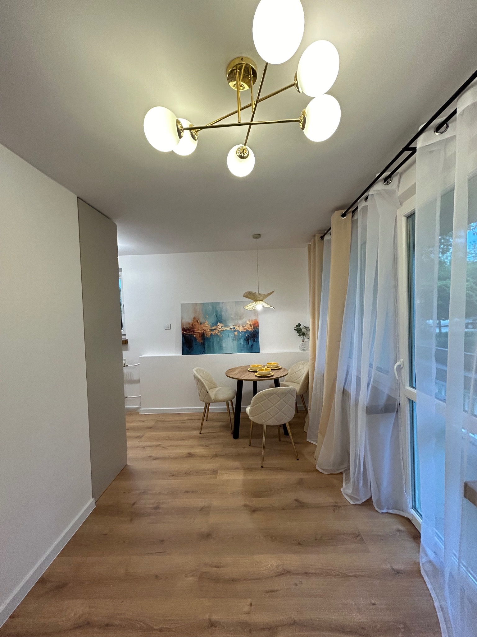 Jasne wnętrze jadalni z okrągłym stołem, krzesłami w stylu glamour, nowoczesną lampą sufitową i abstrakcyjnym obrazem na ścianie. Widoczne drewniane panele podłogowe i zwiewne, białe zasłony.