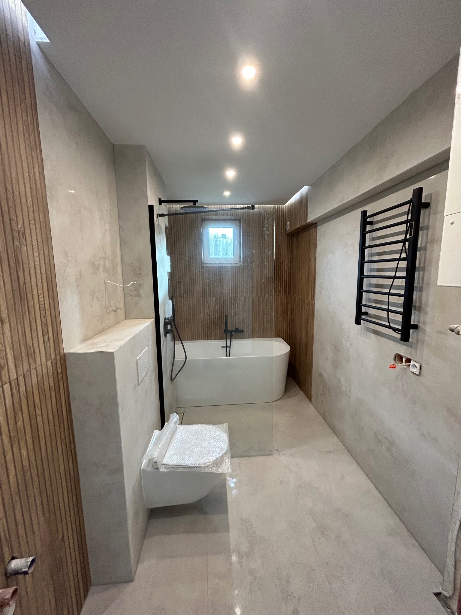 Wykończona łazienka w neutralnych barwach z drewnianymi panelami, wanną wolnostojącą, toaletą w folii ochronnej i grzejnikiem drabinkowym.