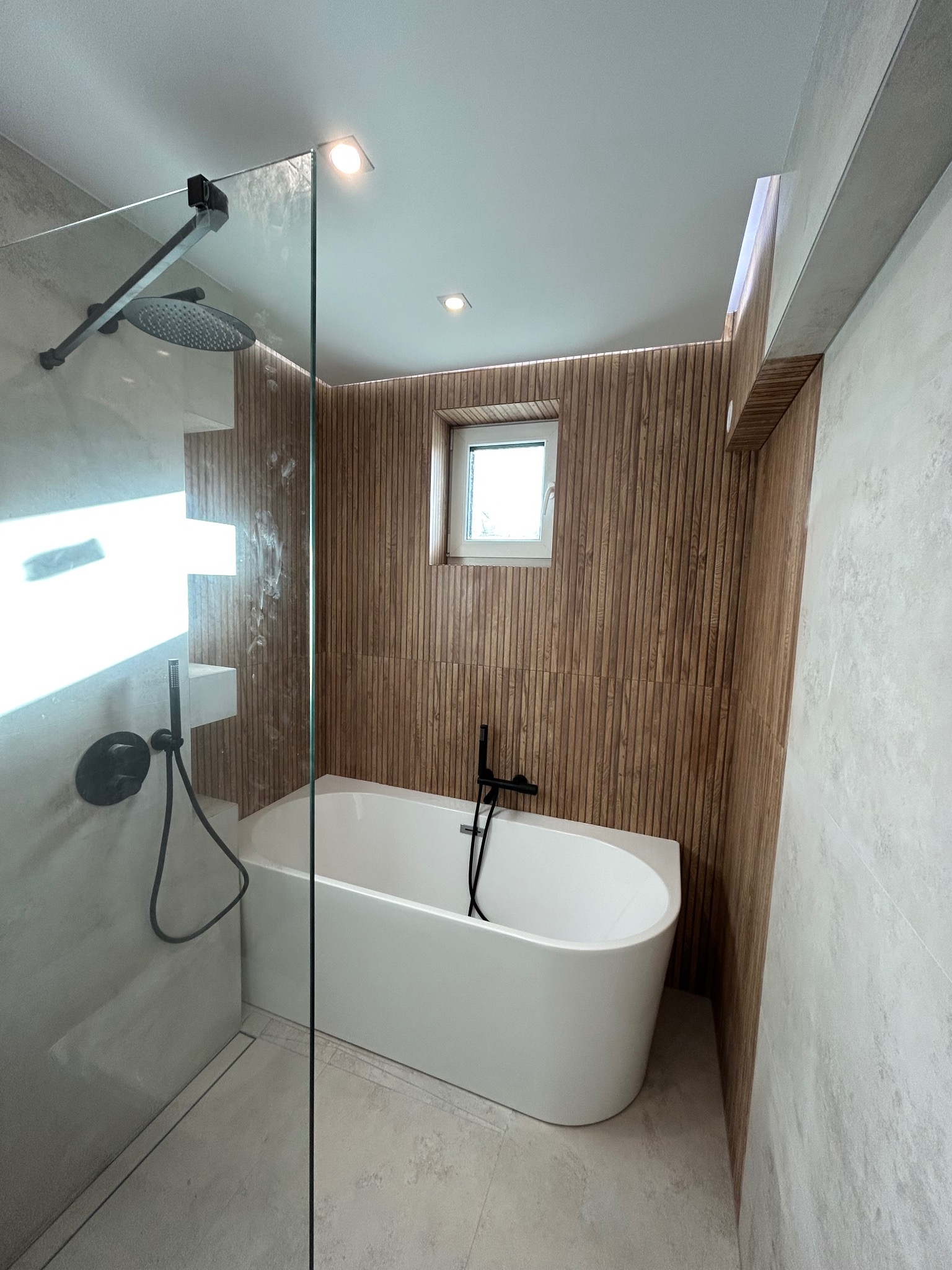 Nowoczesna łazienka z wolnostojącą białą wanną, czarną armaturą i ścianą wykończoną drewnianymi panelami, widok z kabiny prysznicowej, oświetlenie punktowe.