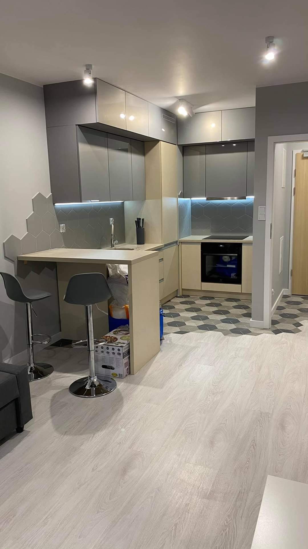 Nowoczesna kuchnia w szaro-beżowej tonacji z wyspą i hokerami, heksagonalne płytki na ścianie i podłodze, oświetlenie LED pod szafkami, widoczne kartony pod wyspą.