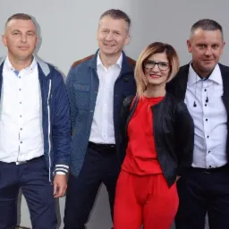 Portret studyjny czteroosobowej grupy w różnym wieku, ubranych w eleganckie, ale nieformalne stroje, stojących na tle jednolitej szarej ściany.