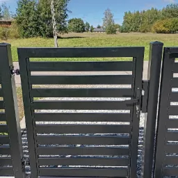 Nowoczesna, czarna furtka z poziomymi panelami na tle zielonego pola i błękitnego nieba, widok z bliska na zamek i klucz.