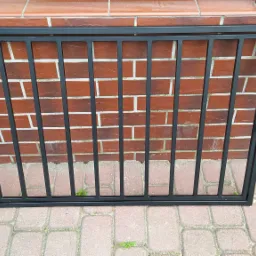 Czarna, metalowa kratka zabezpieczająca okno piwniczne, zamontowana w murze z czerwonej cegły, widoczna na tle chodnika z kostki brukowej.