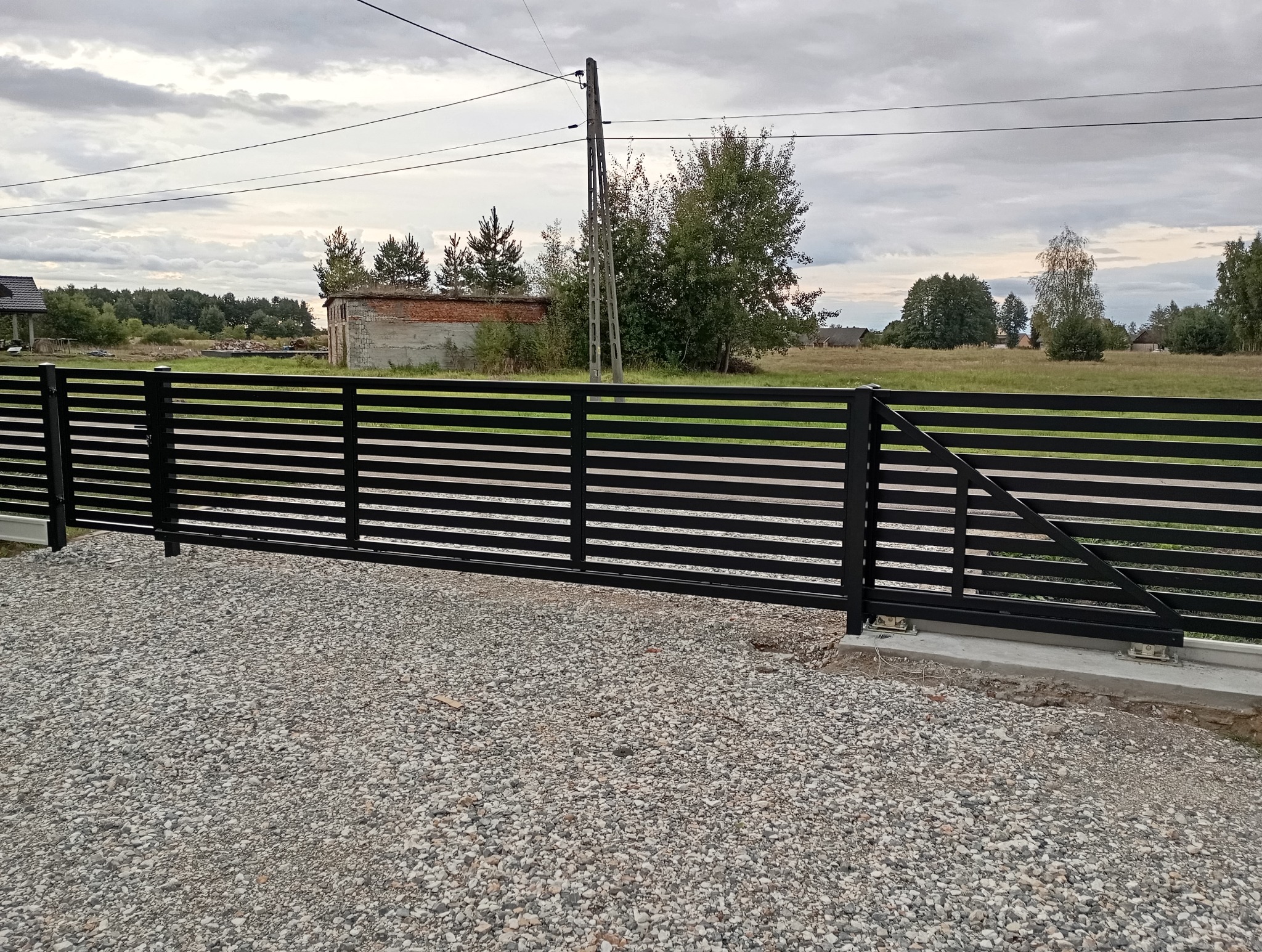 Nowoczesna, przesuwna brama wjazdowa z poziomych, czarnych profili metalowych, zamontowana na betonowej podstawie z widocznymi rolkami, na tle szarego nieba i wiejskiego krajobrazu z budynkiem...
