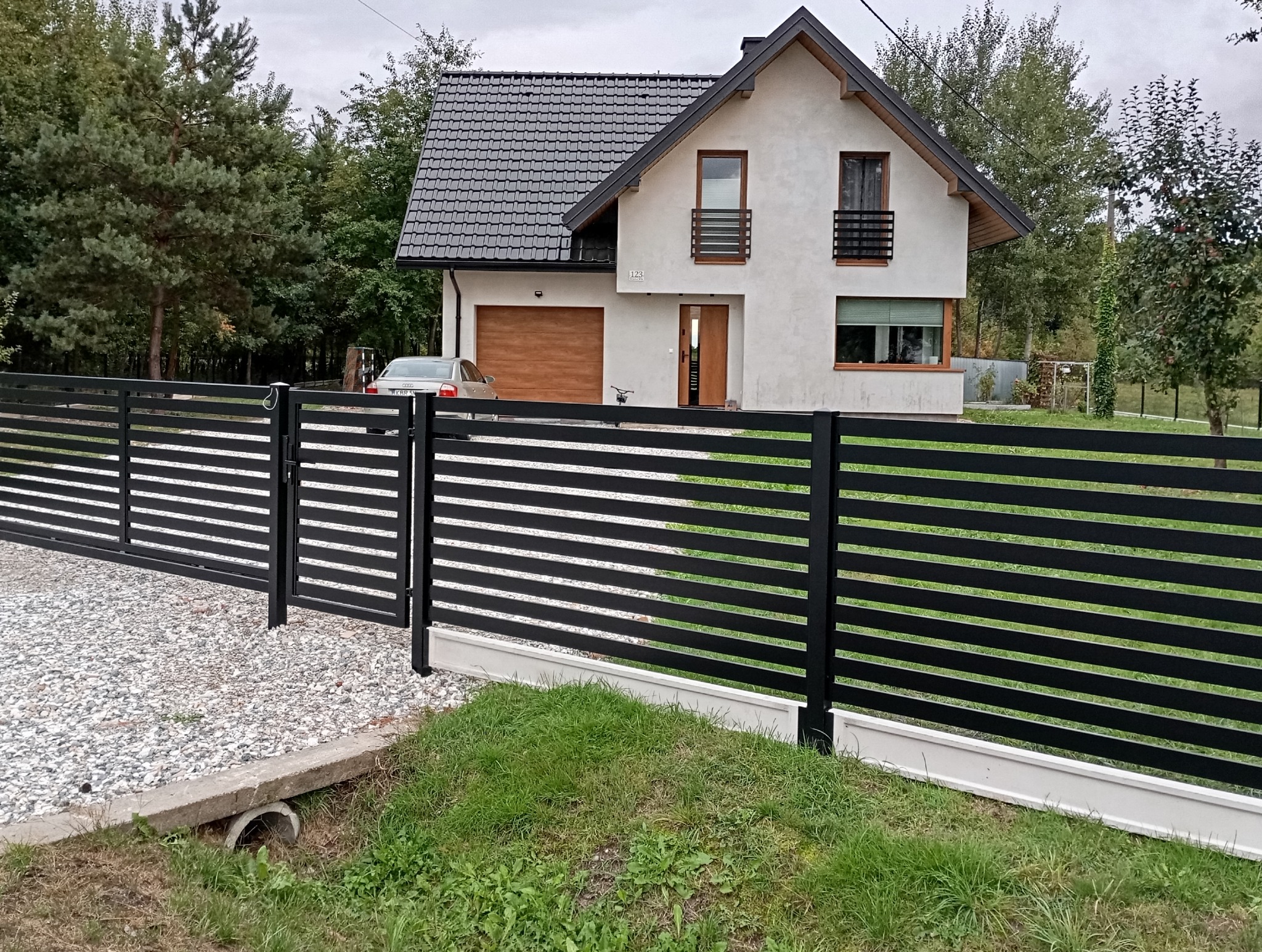 Nowoczesna, pozioma, czarna brama i ogrodzenie z metalowych profili na tle domu jednorodzinnego z garażem i zadbanym trawnikiem.