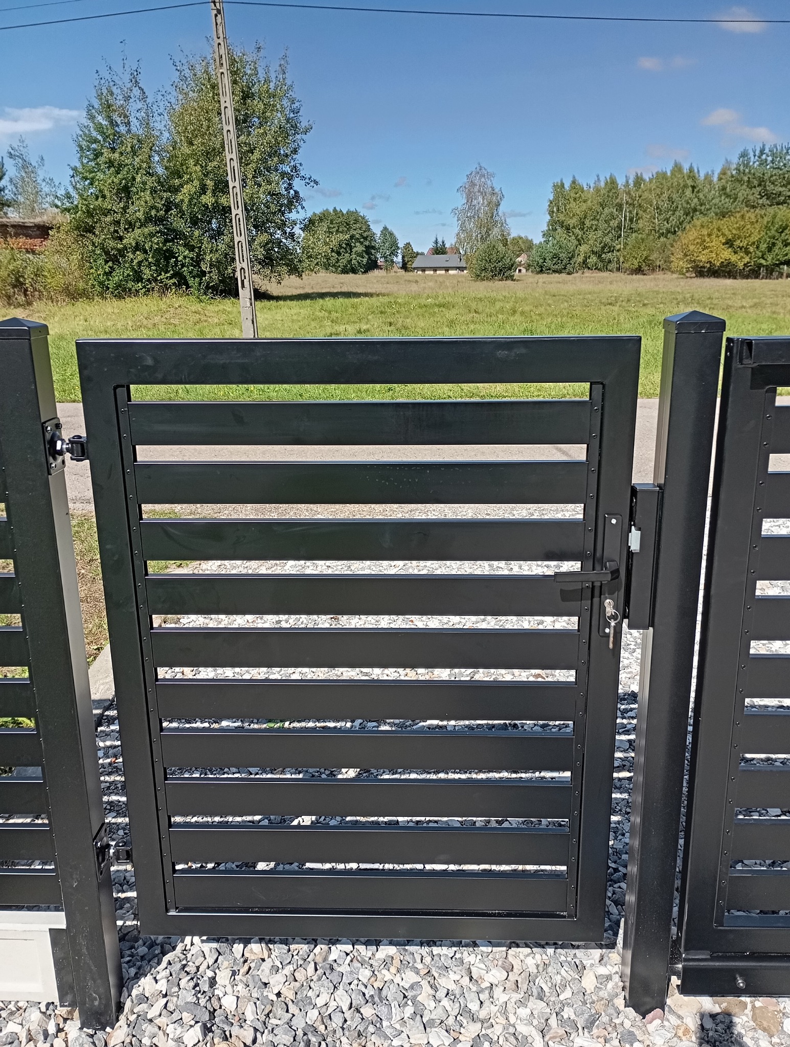 Nowoczesna, czarna furtka z poziomymi panelami na tle zielonego pola i błękitnego nieba, widok z bliska na zamek i klucz.