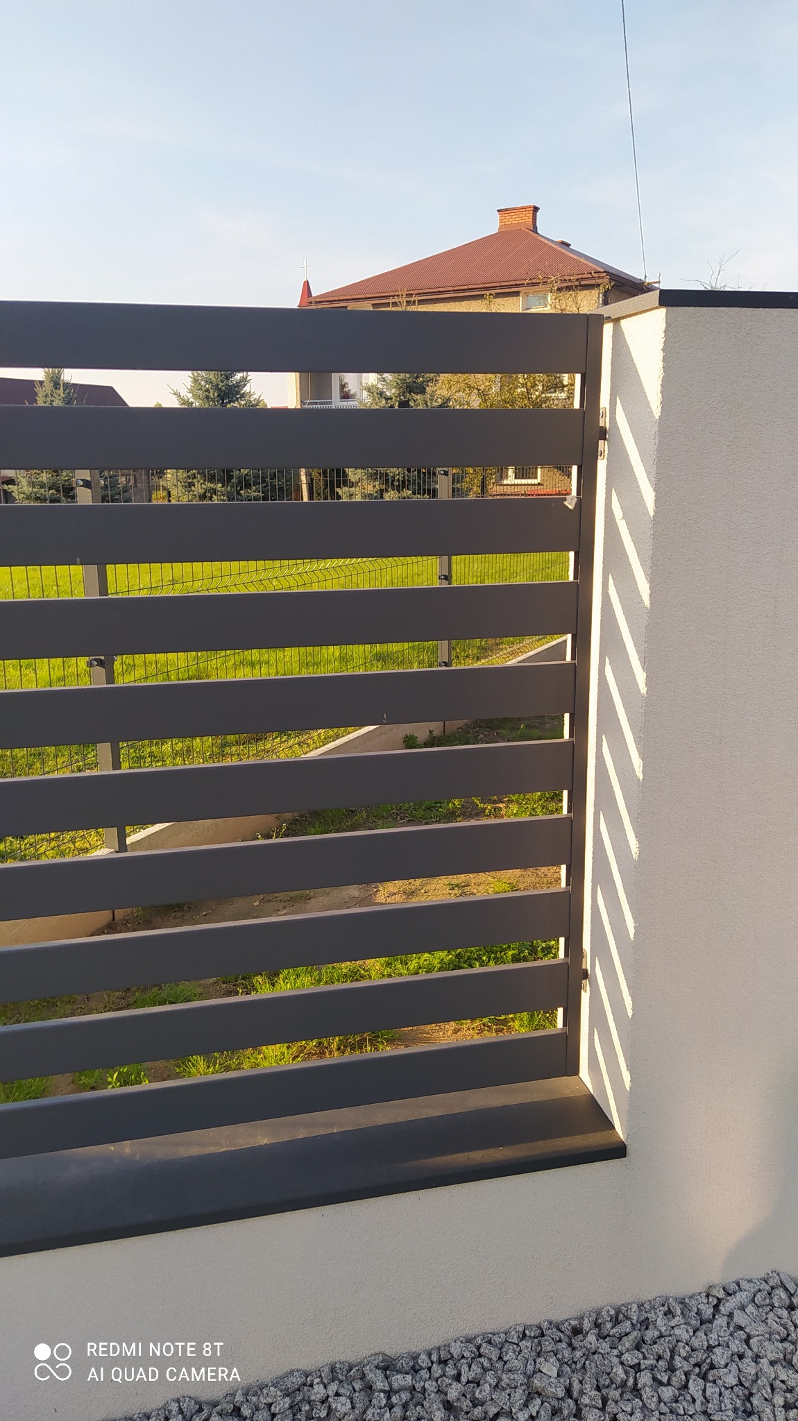 Nowoczesne, poziome ogrodzenie metalowe w kolorze grafitowym, przylegające do białego muru. W tle widoczny budynek z czerwoną dachówką i zielony trawnik.