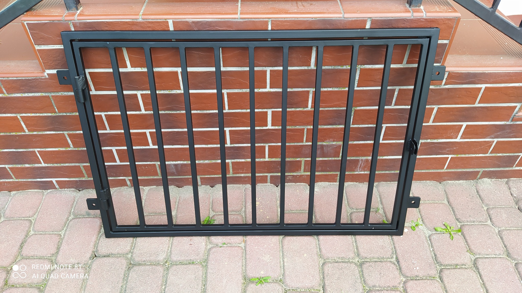 Czarna, metalowa kratka zabezpieczająca okno piwniczne, zamontowana w murze z czerwonej cegły, widoczna na tle chodnika z kostki brukowej.