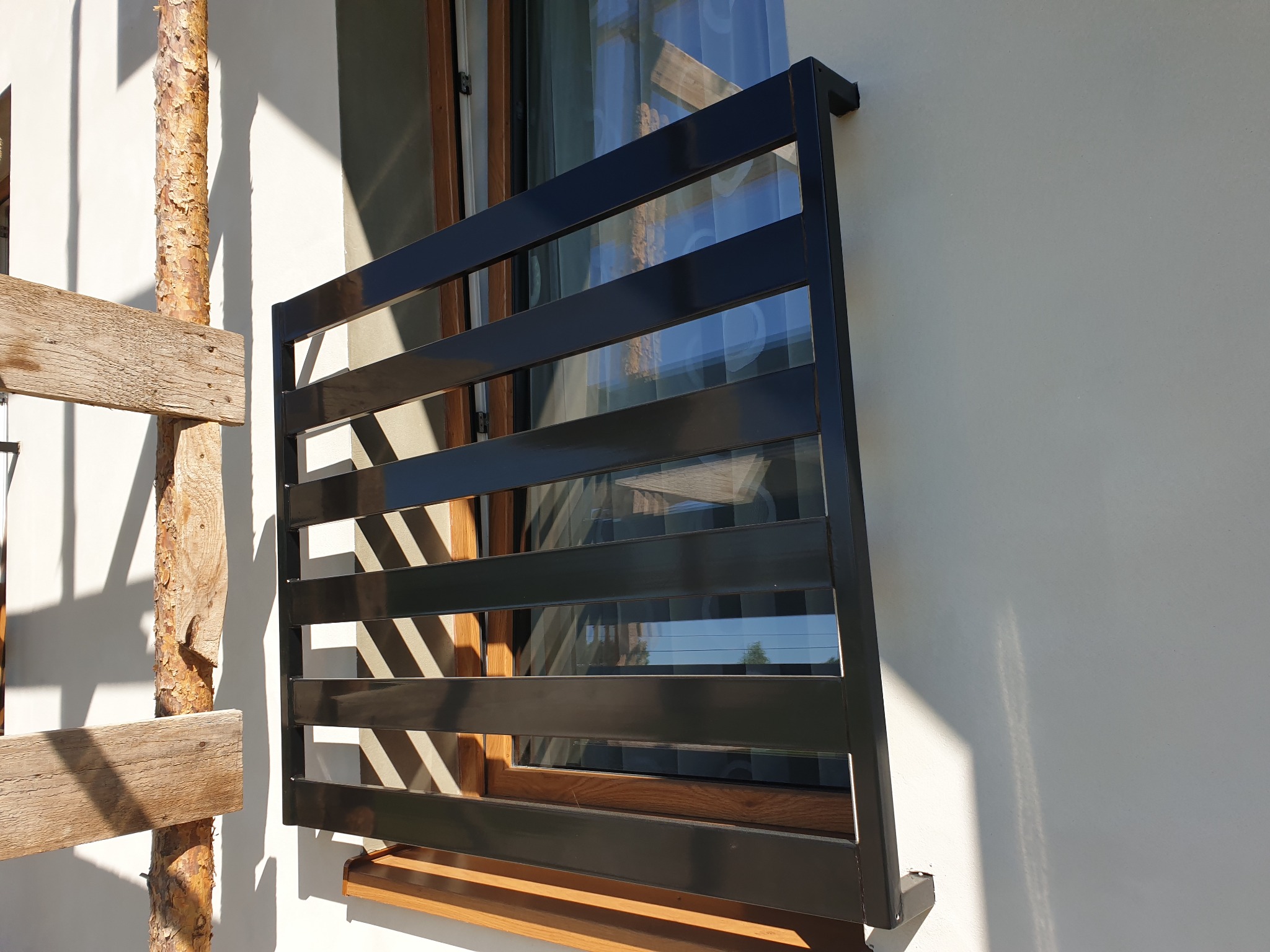 Nowoczesna, czarna, metalowa balustrada balkonowa z poziomych profili, zamontowana przy oknie na tle jasnej elewacji budynku, widoczne drewniane rusztowanie.