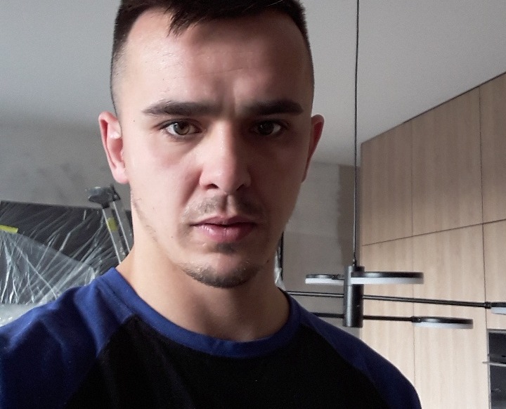 Mężczyzna w trakcie pracy, widoczny fragment kuchni z drewnianymi szafkami i nowoczesną lampą wiszącą, w tle zabezpieczone folią meble.