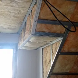 Montaż ocieplenia poddasza wełną mineralną, widoczne metalowe profile konstrukcyjne i przewody elektryczne.