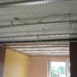 Metalowa konstrukcja sufitu podwieszanego z widocznymi kablami elektrycznymi, częściowo zamontowane płyty sufitowe z fakturą, ściany w różnych kolorach, okno z widokiem na zieleń.