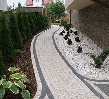 Zakręcający, jasny chodnik z kostki brukowej z ciemnymi obrzeżami, otoczony tujami, korą i białymi kamieniami ozdobnymi, prowadzący wzdłuż ściany budynku.