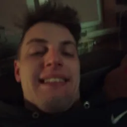 Autoportret uśmiechniętego mężczyzny z bliska, ubrany w ciemną bluzę z widocznym logo Nike, w tle fragment pokoju z oknem.