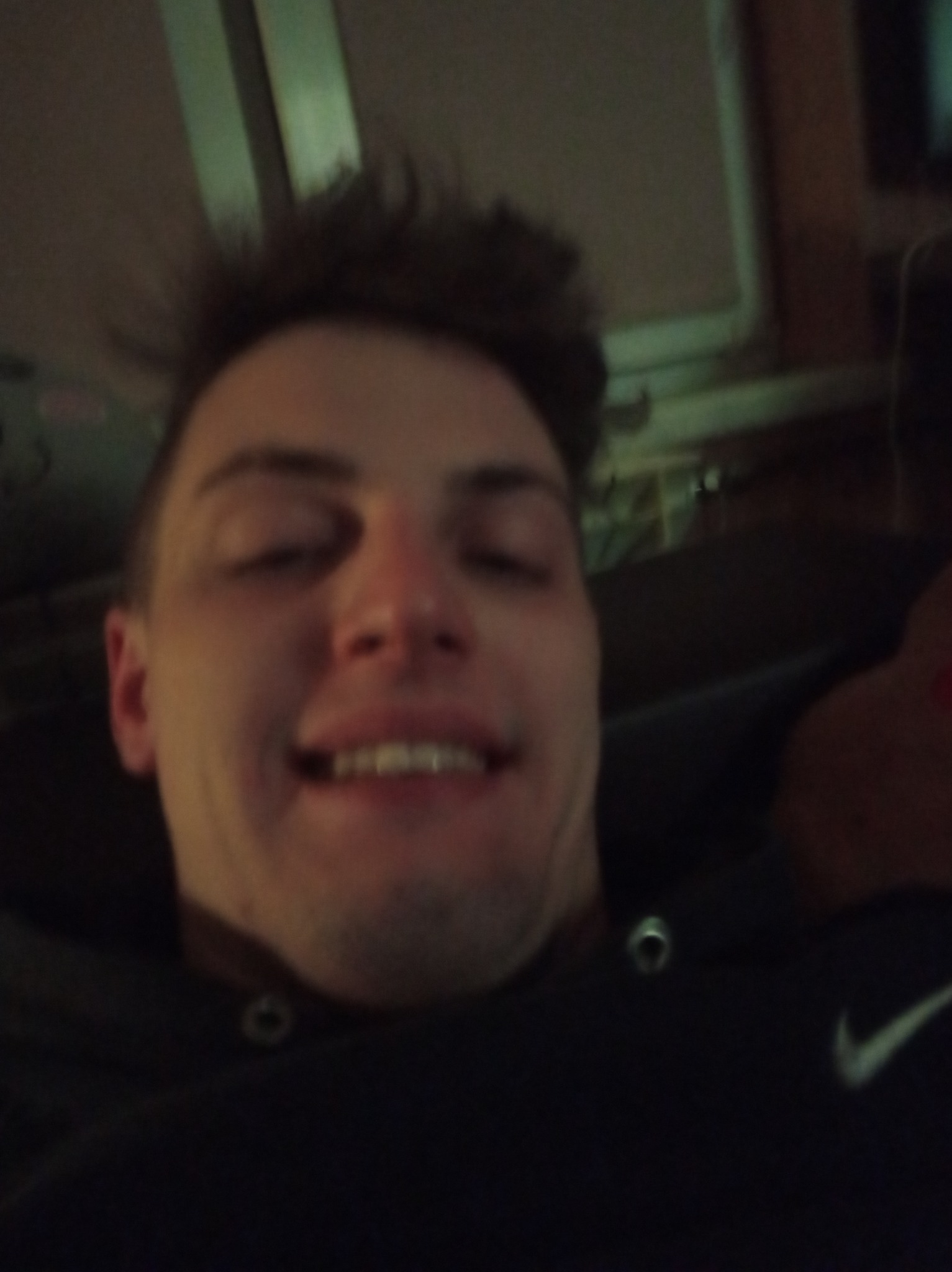 Autoportret uśmiechniętego mężczyzny z bliska, ubrany w ciemną bluzę z widocznym logo Nike, w tle fragment pokoju z oknem.