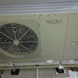 Zewnętrzna jednostka pompy ciepła Netsu z widocznym wentylatorem, częściowo pokryta śniegiem, stojąca na betonowej podstawie w pobliżu ściany budynku i kamiennego podłoża.