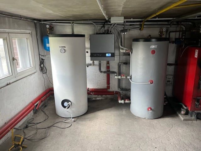 Dwa zbiorniki akumulacyjne ciepła w kotłowni, jeden biały marki Stiebel Eltron, drugi szary z czerwonymi detalami, połączone rurami z systemem sterowania Netsu Premium i kotłem na paliwo stałe.