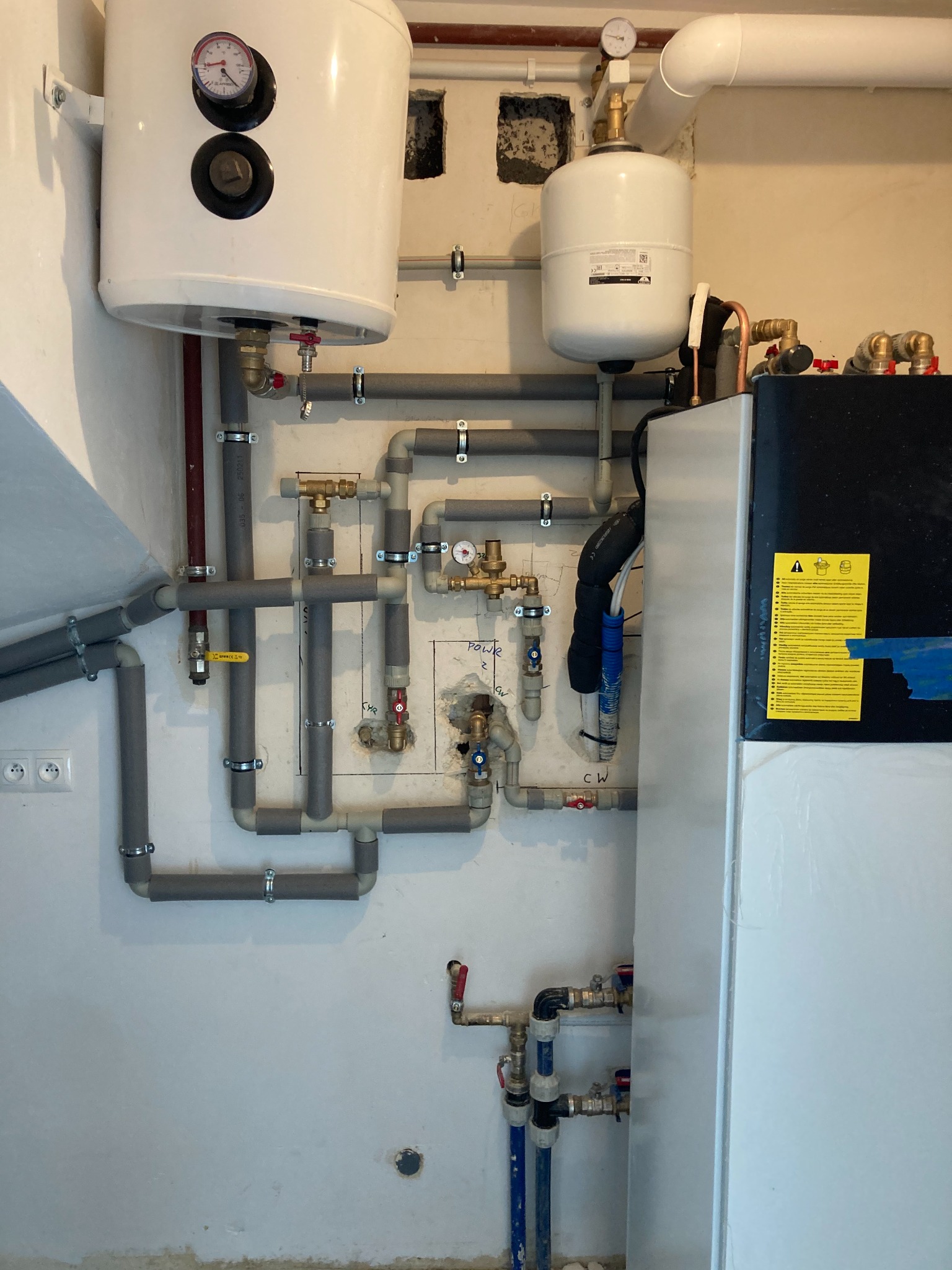 Zintegrowany system grzewczy z zbiornikiem CWU, widoczne rury izolowane termicznie, manometry, zawory, przyłącza elektryczne i schemat instalacji na ścianie.