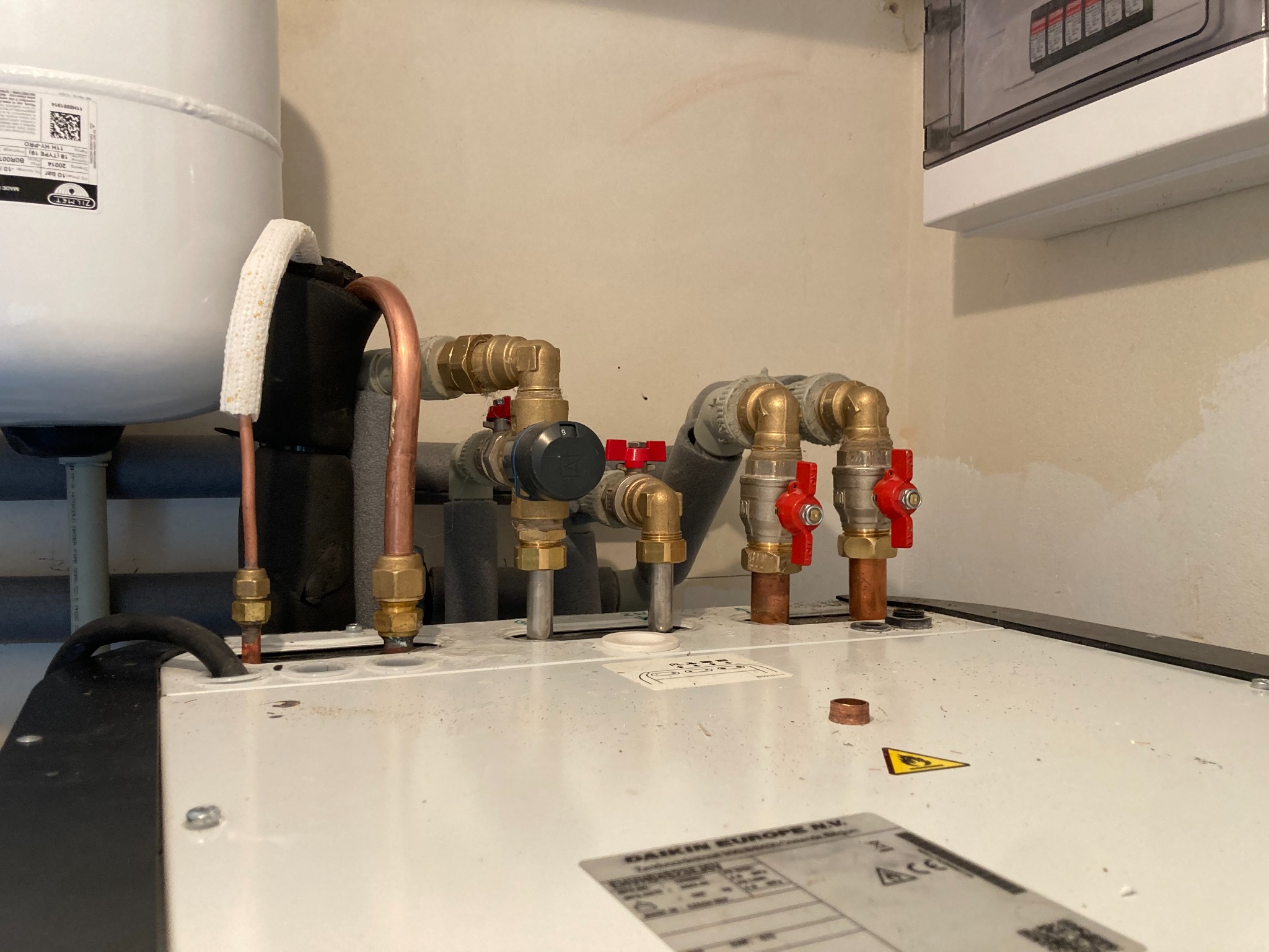 Podłączenie rur miedzianych i zaworów do białego zbiornika, prawdopodobnie elementu pompy ciepła, widoczne etykiety i oznaczenia producenta na urządzeniu, przyłącza zabezpieczone otuliną.